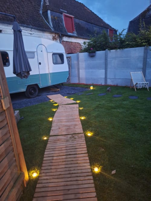 Allée en bois illuminée traversant le jardin luxuriant de la Cour d'Hortense, avec caravane vintage et bâtiments en brique au crépuscule