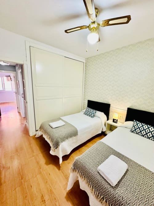 Dormitorio luminoso con tres camas individuales, piso de madera, ventilador de techo y papel tapiz crema en apartamento de Mérida