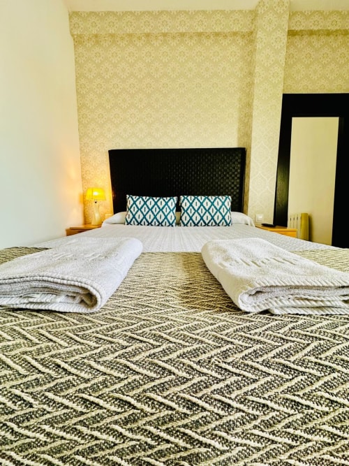 Dormitorio amplio en apartamento de Mérida con toallas dobladas en la cama, alfombra estampada, cabecero negro y almohadas turquesas