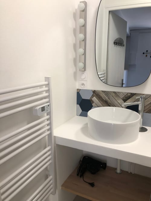 Salle de bain moderne avec lavabo blanc rond, miroir éclairant, porte-serviettes et dosseret en bois à l'appartement Le Boisé