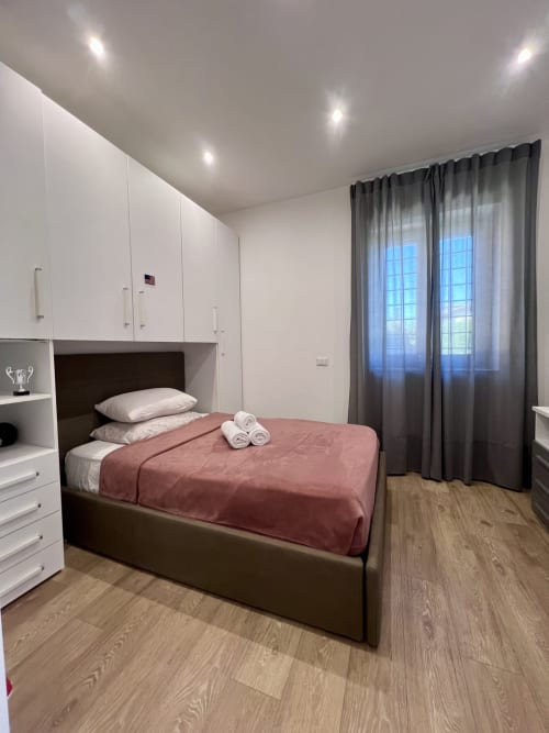 Camera da letto moderna dell'appartamento Zoelia a Roma con letto matrimoniale, armadio bianco, tende grigie, pavimento in legno e vista sulla città