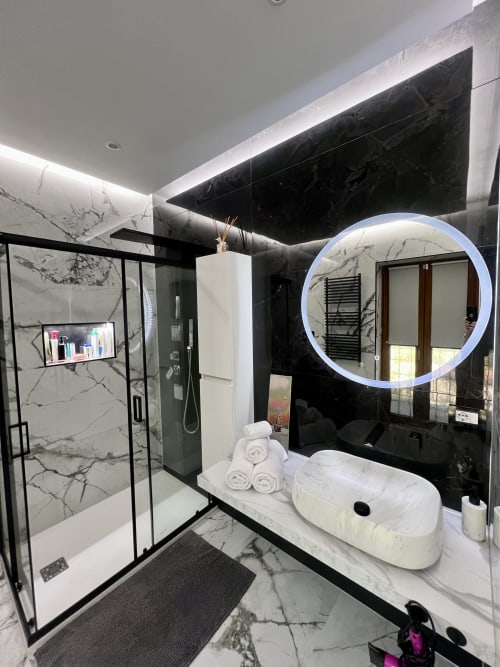 Bagno moderno a Zoelia Roma con pareti in marmo, specchio LED circolare, lavabo bianco sospeso e doccia walk-in