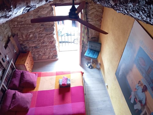 Acogedora habitación en La Calma De Bellver con ropa de cama colorida, paredes de piedra, ventilador de techo y vistas a la montaña desde la puerta