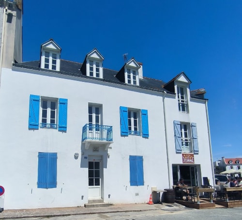 Bâtiment breton blanc aux volets bleus et lucarnes à Locmaria, Belle-Île-en-Mer, avec terrasse de restaurant en bord de mer