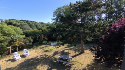 Vue du jardin de la Maison Martha sur Belle Île en Mer avec transats, table de pique-nique et arbres luxuriants