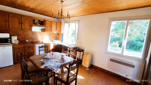 Salle à manger lumineuse avec poutres en bois, cuisine, table ronde et vue sur le jardin à Loc'Maria, Belle Île
