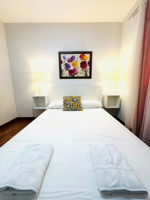 Dormitorio moderno en apartamento Gestyp-AT en Mérida con cama blanca, arte floral colorido, lámparas de mesita y vista al jardín