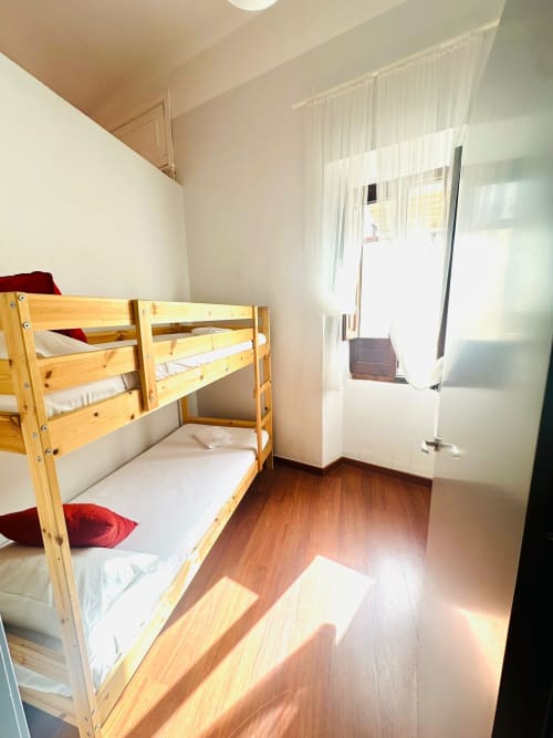 Dormitorio luminoso con literas de madera, ropa de cama blanca, almohadas rojas, piso de parqué y ventana con cortinas translúcidas