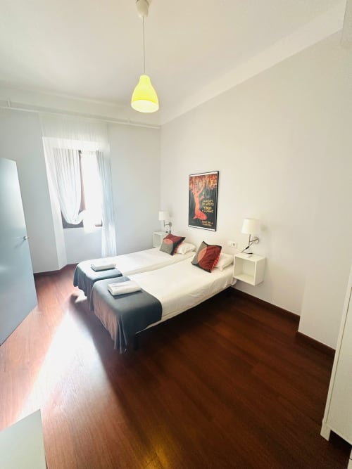 Dormitorio moderno en apartamento de Mérida con camas gemelas, lámpara colgante amarilla, pisos de madera y ventana con vista a monumentos