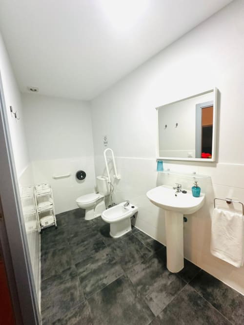 Baño accesible moderno con sanitarios blancos, lavatorio de pedestal, barras de seguridad y piso en cerámica oscura en apartamento Gestyp-AT