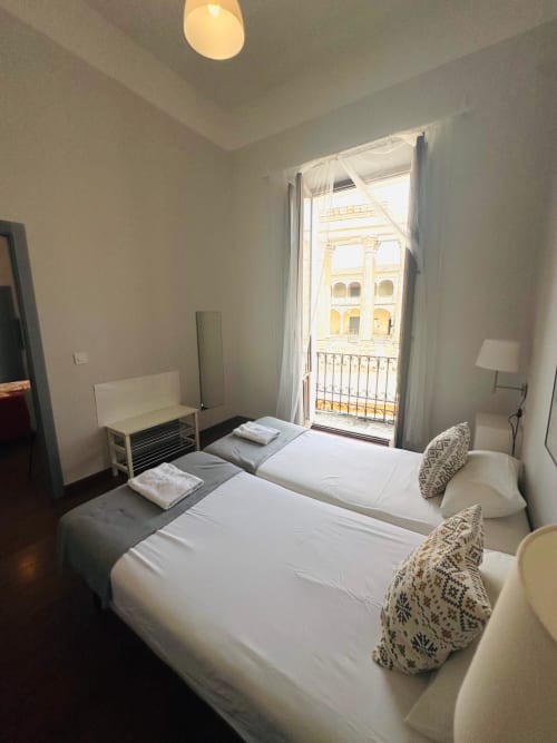 Dormitorio doble con balcón vista a edificio colonial amarillo en Mérida, España. Camas blancas impecables con almohadas decorativas