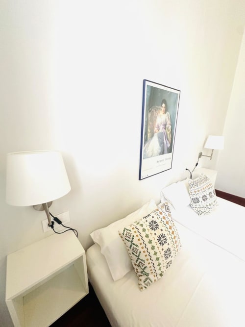 Rincón del dormitorio del apartamento Mérida con mesita de noche blanca, lámpara, almohadas decorativas y cuadro con retrato enmarcado