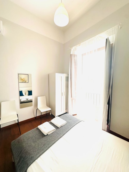 Dormitorio moderno y luminoso en apartamento de Mérida con cama blanca, banco gris, silla, espejo y gran ventana con cortinas