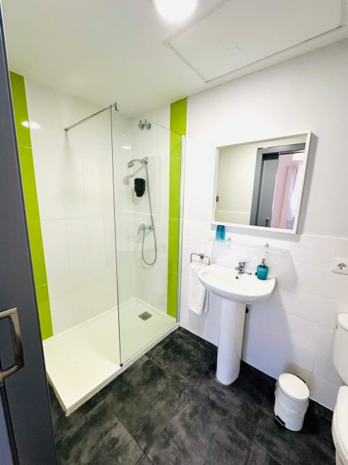 Baño moderno con acentos verde lima, ducha tipo vestidor, lavamanos de pedestal y piso de azulejos oscuros en el apartamento Gestyp-AT