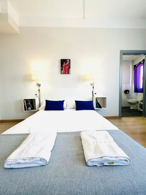 Dormitorio de apartamento moderno en Mérida con cama blanca, almohadas azules, lámparas de pared y baño privado