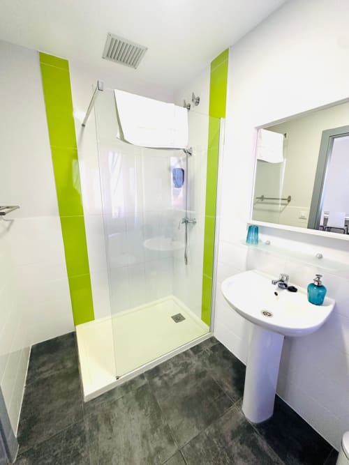 Baño moderno con detalles verde lima, ducha de acceso libre, lavabo pedestal y piso de azulejos oscuros en apartamento Gestyp-AT