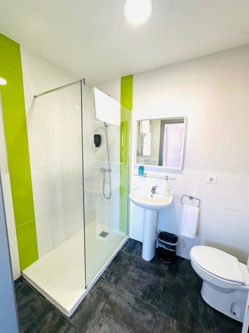 Baño moderno con detalles verde lima, ducha a ras de suelo, lavabo pedestal y piso de azulejos oscuros en apartamento Gestyp-AT