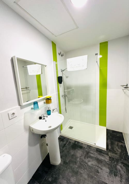 Moderno baño del apartamento Gestyp-AT en Mérida con lavabo blanco, detalles verde limón, ducha amplia y piso de azulejos oscuros.