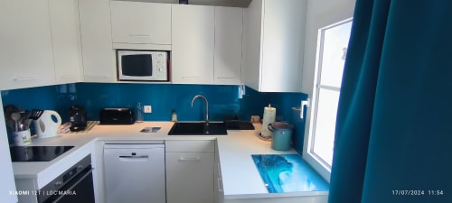 Cuisine moderne blanche avec dosseret turquoise, four à micro-ondes, lave-vaisselle, évier et fenêtre à l'appartement Loc'Maria à Belle-Île-en-Mer