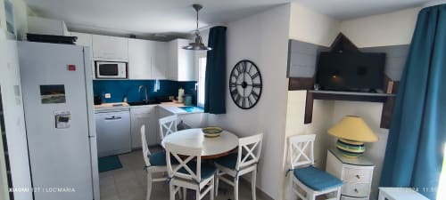 Cuisine-séjour moderne à l'appartement Loc'Maria avec mobilier blanc, dosseret bleu, table ronde, TV et accents turquoise