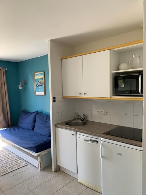 Kitchenette compacte aux armoires blanches, coin détente bleu et mur accent turquoise à Loc'Maria Belle Île en Mer