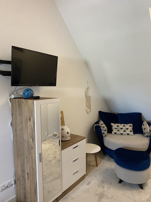 Chambre mansardée moderne aux Chambres d'hôtes Lady Jule : fauteuil bleu marine, commode en bois, téléviseur et décoration macramé