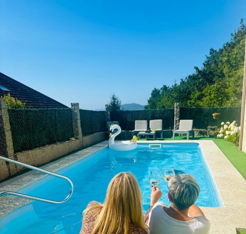 Dos huéspedes disfrutan del vino junto a la piscina azul de la casa rural Casa Caeiro en Baiona, con vistas a las montañas y tumbonas verdes en el jardín.