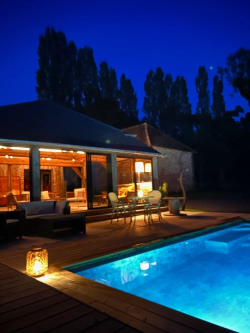 Domaine de Blanvatel illuminated at dusk with glowing pool, wooden deck, and tall cypress trees silhouetted against blue sky.