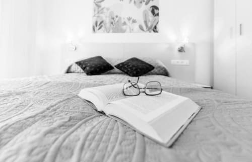 Dormitorio minimalista en apartamento Gestyp-AT Mérida con libro abierto y gafas sobre la cama, almohadas negras y arte botánico