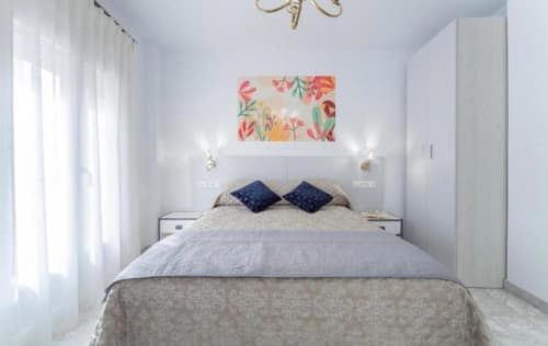 Luminoso dormitorio moderno en apartamento de Mérida con cama doble, almohadas azules, obra floral y vistas a la ciudad