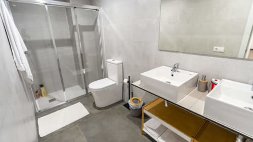 Baño moderno en Apartamentos Burriana con doble lavamanos, ducha, inodoro y mueble de almacenamiento de madera.