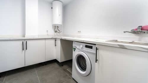 Moderno cuarto de lavado en Apartamentos Burriana con lavadora blanca, calentador de agua y armarios de almacenamiento
