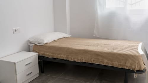 Luminosa habitación en Apartamentos Burriana con cama individual, almohada blanca, manta beige, mesita de noche y ventana con cortina translúcida