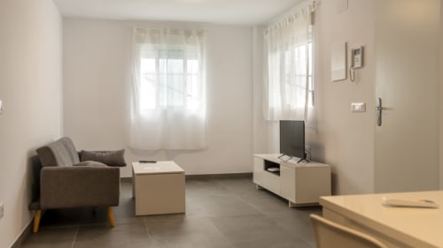 Luminoso salón de Apartamentos Burriana con sofá gris, muebles blancos y luz natural de las ventanas