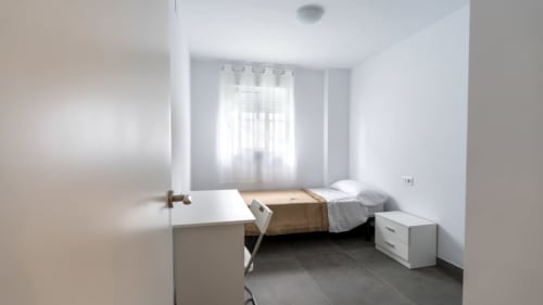 Dormitorio luminoso en Apartamentos Burriana con cama individual, escritorio, mesita de noche y gran ventana con cortinas translúcidas