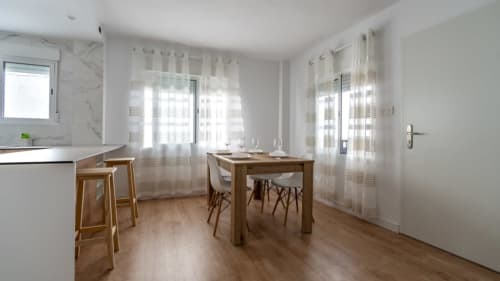 Área moderna de cocina-comedor en Apartamentos Burriana con mesa de madera, barra de desayuno y ventanas luminosas