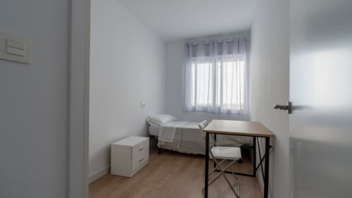 Luminoso dormitorio en Apartamentos Burriana con cama individual, escritorio, ventana con cortinas translúcidas y suelo de madera.