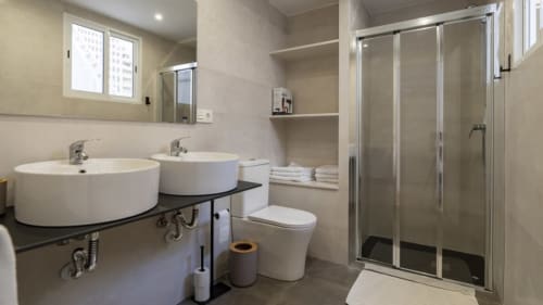 Baño moderno en Apartamentos Burriana con dos lavabos redondos, inodoro y ducha con plato de obra con puertas de cristal