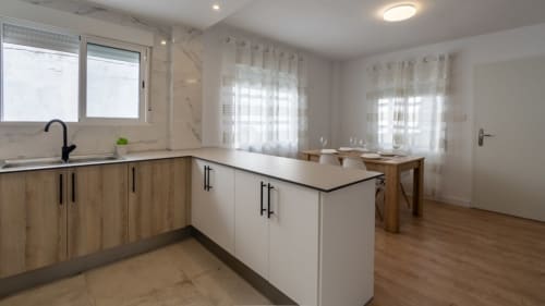 Cocina y comedor moderno en apartamento Burriana con muebles blancos, detalles de madera y ventanas luminosas.