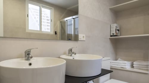 Baño moderno en Apartamentos Burriana con dos lavabos redondos, espejo, estantería y vistas a la ciudad a través de la ventana