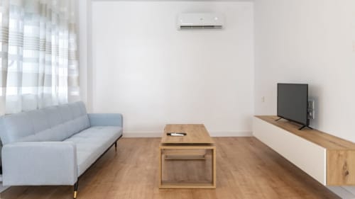Moderna sala de estar en Apartamentos Burriana con sofá gris, mesa de madera, televisor y grandes ventanales iluminados