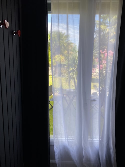Des rideaux blancs aérés encadrent la vue d'un jardin ensoleillé avec palmiers et fleurs roses aux Chambres d'hôtes Lady Jule