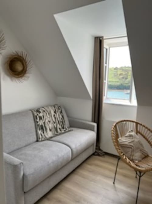 Chambre lumineuse sous les toits avec canapé gris, fauteuil en rotin et vue mer à Locmaria Belle Île en Mer
