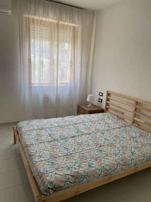 Camera da letto luminosa con letto in legno, biancheria patterned, tende trasparenti e vista mare a Rigiariloo Home Apartments a San Giacomo