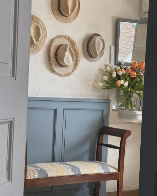 Élégante entrée à La Maison De Fleurat avec chapeaux tressés sur mur crème, lambris bleu, chaise en bois et fleurs fraîches