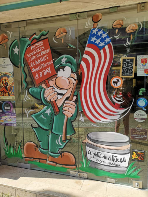 Colorful storefront mural at Au Gite d'Isigny featuring cartoon leprechaun, American flag, and French pâté jar in Normandy
