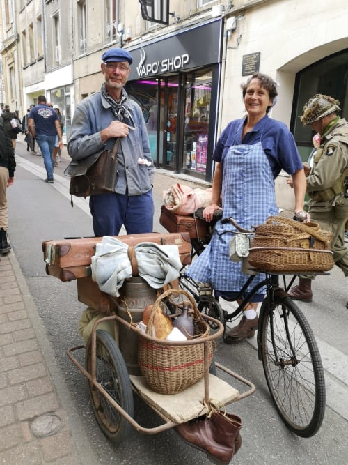 Deux personnes posant avec un vélo vintage et des marchandises dans une rue d'Isigny-sur-Mer, France