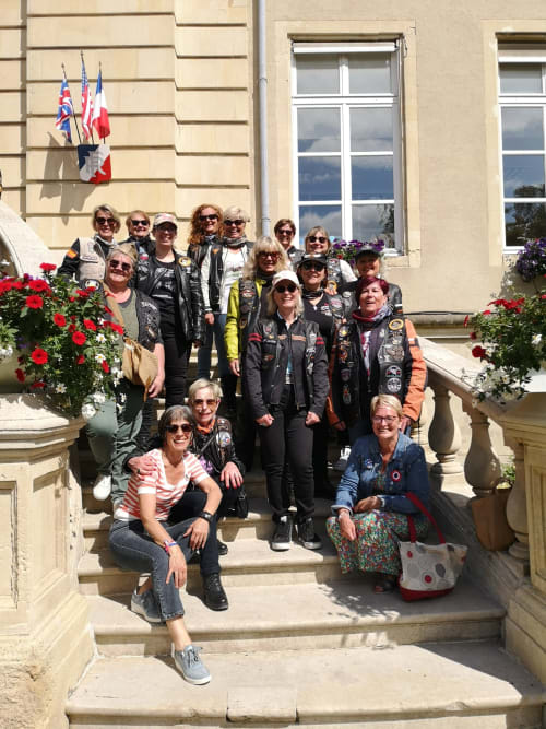 Group of women bikers posing on steps at Au Gite d'Isigny B&B in Isigny-sur-Mer, France with flags and flowers