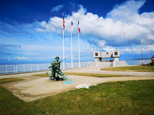 D-Day memorial statue at Isigny-sur-Mer waterfront with flags and monument overlooking Normandy coast