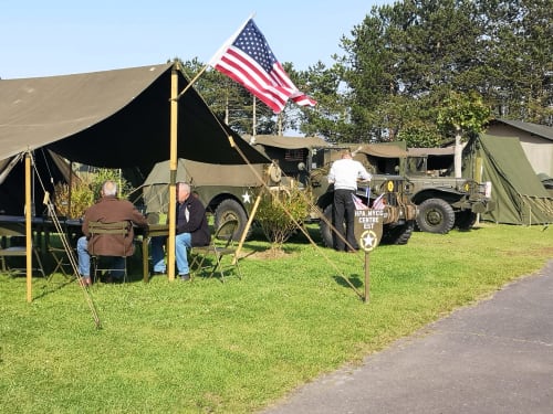 WWII military encampment display with American flag, vintage trucks, and canvas tents at Isigny-sur-Mer historical event